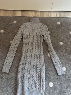 Grå stickad poloklänning - Säljer en lång, grå stickad klänning med polokrage och kabelstickat mönster. Klänningen har långa ärmar och ribbade muddar vid ärmslut och nederkant. Perfekt för kyliga dagar och enkel att styla med boots eller sneakers. Har ett litet hår i halsen där bak så priset går att diskuteras.