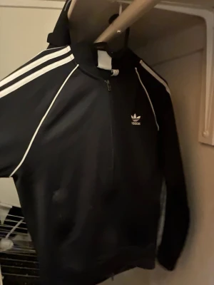 Svart Adidas hoodie med dragkedja - Säljer en svart hoodie från Adidas med klassiska vita ränder längs ärmarna och vit Adidas-logga på bröstet. Tröjan har dragkedja framtill, huva och är tillverkad i ett mjukt material som passar perfekt till en sportig stil.
