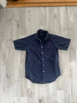 Mörkblå skjorta från Ralph Lauren - Klassisk mörkblå skjorta från Ralph Lauren med korta ärmar och knappar framtill. Skjortan har den ikoniska röda logotypen broderad på bröstet och är tillverkad i mjuk bomull. Perfekt för en avslappnad men ändå stilren look.