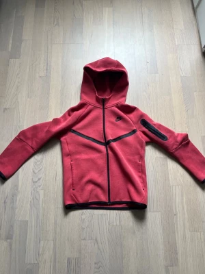 Röd Nike hoodie med svarta detaljer - Säljer en röd hoodie från Nike med svarta dragkedjor och detaljer. Hoodien har hel dragkedja framtill, fickor på sidorna och en extra ficka på ena ärmen. Tillverkad i ett mjukt och stretchigt material, perfekt för en sportig och avslappnad stil.