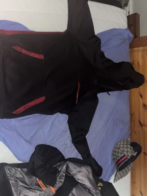Svart och röd hoodie från Prada - Säljer en svart hoodie från Prada med röda detaljer och stor PRADA MILANO-logga på ryggen. Hoodien har huva och känguruficka framtill med röda mesh-liknande inslag. Materialet ser ut att vara en mix av bomull och polyester.