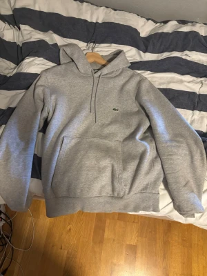 Grå hoodie från Lacoste - Snygg grå hoodie från Lacoste med klassisk krokodillogga på bröstet. Hoodien har huva med dragsko och en stor magficka framtill. Perfekt för en chill och stilren look. Materialet är mjukt och skönt,  bomullsblandning.