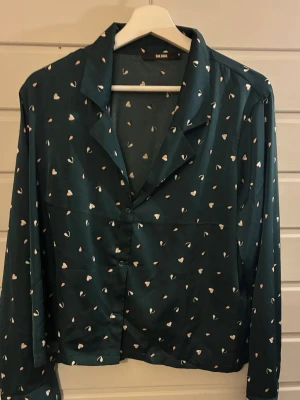 Mörkgrön blus med hjärtmönster - Snygg mörkgrön blus från Bik Bok med små vita hjärtan över hela tyget. Blusen har klassisk krage, knäppning framtill och långa ärmar. Materialet är tunt och ger ett mjukt, flowy intryck. Perfekt för dig som gillar söta detaljer och en avslappnad look.