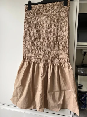 Beige volangkjol med smock - Supersöt beige kjol med smock upptill och volang nedtill. Kjolen har en lätt glansig finish och är perfekt för dig som gillar en feminin och trendig stil. Går att ha både som miniklänning o kjol, se produktbilderna. 