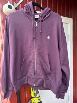 Lila hoodie med dragkedja från Carhartt stl L (w) - Snygg lila hoodie från Carhartt med dragkedja framtill, huva med dragsko och känguruficka. Liten vit Carhartt-logga på bröstet. Perfekt för en avslappnad streetwear-look. Tillverkad i mjuk bomullsmix för extra komfort. Boxig modell. Bröstvidd 55. Plagglängd 60