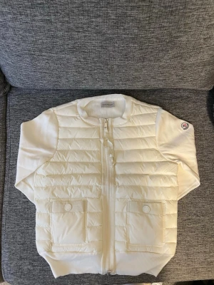 Vit dunväst från Moncler - Snygg vit dunväst från Moncler med quiltad framsida, ribbade detaljer och två fickor med knapp framtill. Västen har dragkedja och Moncler-logga på ärmen. Perfekt för lager-på-lager och ger en clean look till din outfit.Storleken är 1 vilket motsvarar S