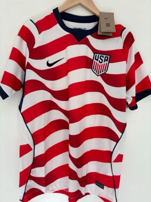 USA Nike fotbollströja röd/vit - Snygg fotbollströja från Nike med USA-märke på bröstet. Tröjan har röda och vita vågiga ränder, blå detaljer vid krage och ärmslut samt Dri-FIT-material som andas. Perfekt för träning eller match och har en sportig, modern look.