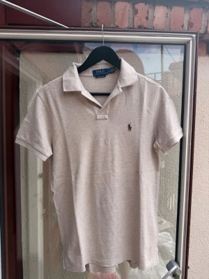 Beige pikétröja från Polo Ralph Lauren - Klassisk beige pikétröja från Polo Ralph Lauren med korta ärmar och krage. Tröjan har den ikoniska broderade loggan på bröstet och är tillverkad i mjuk bomull. Perfekt till en avslappnad men ändå stilren look.