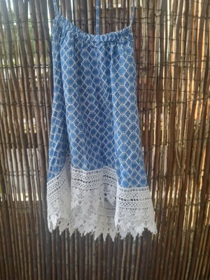 Blå mönstrad topp med spets - Söt blå topp med vitt geometriskt mönster och tunna axelband. Nederdelen har bred vit spetskant med blommiga detaljer som ger en bohemisk känsla. Perfekt för varma dagar och sommarens alla äventyr. Katt finns i hemmet.