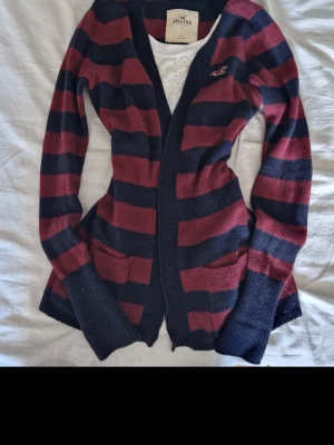 Vintage kardigan hollister stripes y2k 2000s alternative emo scene fall dark coquette  - Mysig kofta från Hollister med breda marinblå och vinröda ränder. Koftan har två fickor framtill, ribbade muddar och en liten broderad logga på bröstet. Perfekt för lager på lager och en avslappnad stil.