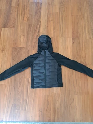 Svart vindjacka med avtagbar huva - Snygg svart vindjacka med avtagbar luva och dragkedja framtill. Jackan har släta ärmar i stretchigt material och vadderad front. Praktiska sidofickor med dragkedja. Perfekt för blåsiga dagar och enkel att matcha med olika outfits.😍