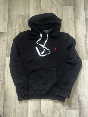 Svart hoodie från Polo Ralph Lauren - Svart hoodie från Polo Ralph Lauren med vit dragsko och röd broderad logga på bröstet. Klassisk känguruficka framtill och ribbade muddar vid ärmar och midja. Tillverkad i mjukt bomullsmaterial för en skön känsla.