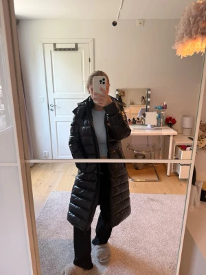 Svart lång dunjacka från Moncler - Säljer en svart, glansig lång dunjacka från Moncler med huva och dragkedja framtill. Jackan har Moncler-logga på ärmen och en figurnära passform som ger en snygg siluett. Perfekt för kalla vinterdagar och riktigt varm.
