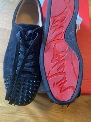 Svarta sneakers med nitar - Säljer ett par svarta sneakers med mattsvarta nitar på tån och klassisk röd sula. Skorna är i mocka och har svarta skosnören. Perfekta för dig som vill sticka ut med en edgy och lyxig stil.