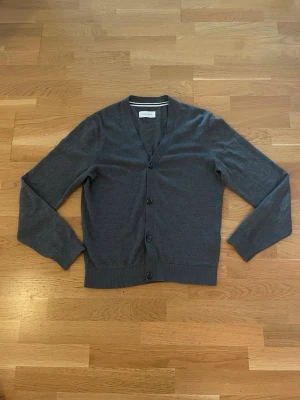 Grå cardigan från Pier One - Stilren grå cardigan från Pier One med v-ringning och fyra knappar framtill. Tillverkad i mjukt, tunt stickat material och har långa ärmar samt ribbade muddar vid ärmslut och nederkant. Perfekt att slänga över en t-shirt eller skjorta.