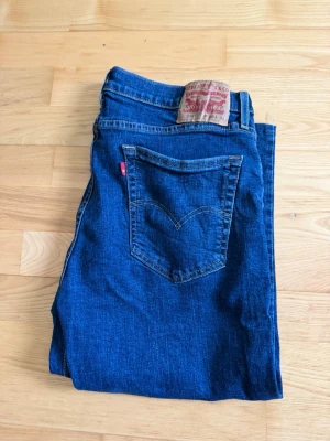 Levi's 514 blå raka jeans - Klassiska blå Levi's 514 jeans med rak passform och femficksdesign. Jeansen har tydliga kontrastsömmar, läderpatch bak i midjan och den ikoniska röda Levi's-taggen på bakfickan. Tillverkade i slitstark denim, perfekta för en avslappnad och tidlös stil.kortare en vanliga jeans, perfekt för sommaren 