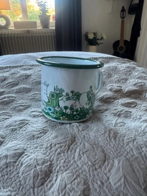 Mumin emaljmugg med grönt motiv - Säljer en vit emaljmugg från Muurla med grönt kantband och motiv av Muminfamiljen bland blommor och växter. Muggen har ett handtag och är dekorerad med detaljerade illustrationer i grönt och vitt. Perfekt för te, kaffe eller varm choklad. Rymmer 3,7 dl och är helt oanvänd
