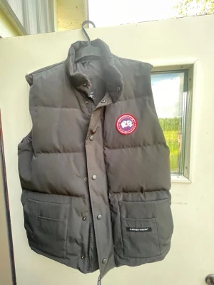 Svart dunväst från Canada Goose - Snygg svart dunväst från Canada Goose med hög krage och tryckknappar. Västen har två stora framfickor och det klassiska runda Canada Goose-märket på bröstet. Perfekt för kyliga dagar och enkel att matcha med olika outfits.