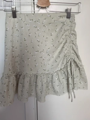 Blommig kortkjol med volang och snörning - Supersöt ljus kortkjol med små blommor, volangkant och snörning på sidan. Kjolen har en feminin och trendig look, perfekt för varma dagar. Materialet känns lätt och luftigt, och mönstret ger en romantisk vibe.