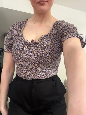 Blommig croppad blus med puffärm - Söt croppad blus med blommigt mönster i blått, rosa, gult och vitt. Toppen har korta puffärmar och smockad rygg för skön passform. Perfekt att styla med höga jeans eller kjol. Har bygel under bröst och mage som sitter skönt