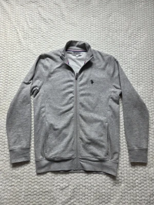 Grå ziptröja från U.S. Polo Assn. - Säljer en grå sweater med fullzip från U.S. Polo Assn Med broderad logga på bröstet. Tröjan har en ståkrage med röd och marinblå randig detalj på insidan samt två fickor framtill. Den har använts en gång och är som sprillans ny! Givetvis äkta! Hör av er vid frågor!