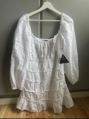 Vit broderad kortklänning med volanger - Supersöt vit kortklänning med broderade detaljer och volanger. Klänningen har långa puffärmar, fyrkantig ringning och är gjord i luftig bomull. Perfekt för sommarens alla tillfällen och ger en romantisk vibe. Den är helt ny och aldrig använd
