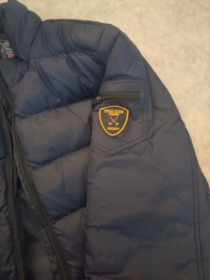 Mörkblå dunjacka Polo Club Vinson - Snygg mörkblå dunjacka från Polo Club Vinson med quiltade detaljer och dragkedjor på fickorna. Jackan har en patch på ärmen och ståkrage för extra värme. Perfekt för kalla vinterdagar och har en sportig, modern look.