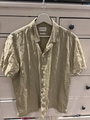 Beige linneskjorta från Lindbergh - Säljer en beige kortärmad skjorta från Lindbergh White i relaxed fit. Skjortan är tillverkad i linne och har klassisk krage samt knappar framtill. Perfekt för varma dagar och enkel att styla till olika outfits. Den är i fint skick men behöver strykas vilket jag kan göra innan jag fraktar den. 
