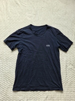 Mörkblå Hugo Boss t-shirt - Snygg mörkblå t-shirt från Hugo Boss med diskret logga broderad på bröstet. Klassisk rund halsringning och korta ärmar. Tillverkad i mjuk bomull som känns skön mot huden. Perfekt till jeans eller shorts för en clean och enkel stil. Priset kan självklart diskuteras!