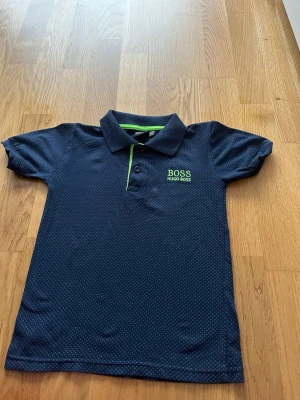 Mörkblå polotopp från Hugo Boss - Snygg mörkblå polotopp från Hugo Boss med diskret rutig mönster och grön broderad logga på bröstet. Kragen har en grön detalj och toppen är kortärmad, perfekt för en cool och avslappnad look.
