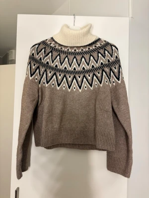 Beige mönstrad polotröja från H&M - Stickad beige polotröja från H&M med grafiskt mönster i svart och vitt över axlar och bröst. Tröjan har hög polokrage, långa ärmar och ribbade muddar. Perfekt för kalla dagar och ger en trendig look.