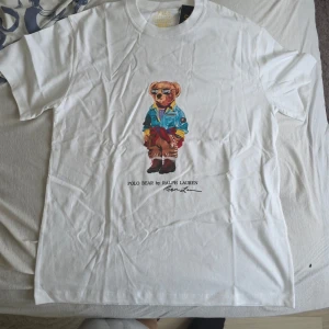 Vit t-shirt Polo Bear Ralph Lauren - Vit t-shirt från Ralph Lauren med ikoniskt Polo Bear-tryck framtill. Klassisk passform och rund halsringning. Perfekt för en avslappnad och trendig stil.