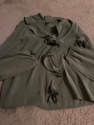 Olivgrön blus med knytning - Olivgrön blus från Vero Moda med långärmad design och dekorativa knytband framtill. Blusen har en bred krage och är tillverkad i ett mjukt, tunt material som ger en avslappnad känsla. Perfekt för dig som gillar en enkel men cool stil.