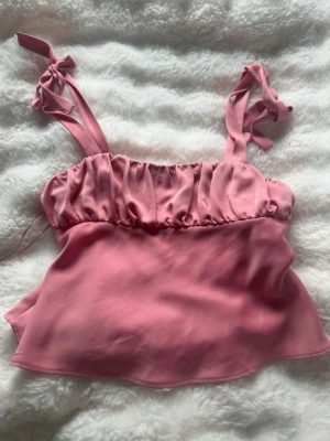 Rosa linnetopp med knytband - Supersöt rosa linnetopp med knytbara axelband och rynkad överdel. Toppen har en croppad passform och är tillverkad i ett mjukt, glansigt material som ger en lyxig känsla. Perfekt att styla med jeans eller kjol för en trendig look.