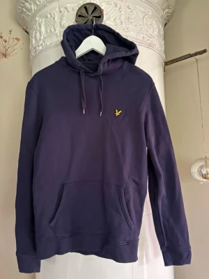 Blå hoodie från Lyle & Scott - Snygg blå hoodie från Lyle & Scott med klassisk gul logga på bröstet. Hoodien har huva med dragsko, känguruficka framtill och ribbade muddar vid ärmar och nederkant. Tillverkad i mjuk bomullsblandning, perfekt för en avslappnad stil.