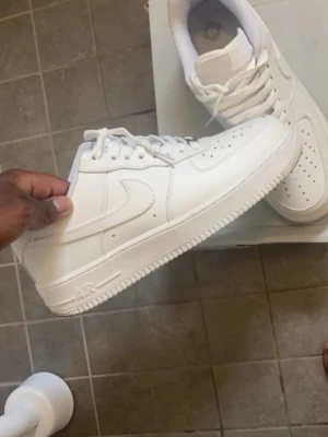 Nike Air Force 1 vita sneakers - Klassiska helvita Nike Air Force 1 sneakers i lågt utförande. Skorna har en stilren design med perforerad tå, vita skosnören och den ikoniska swoosh-loggan på sidan. Tillverkade i slätt läder med en robust gummisula för extra komfort och streetkänsla.
