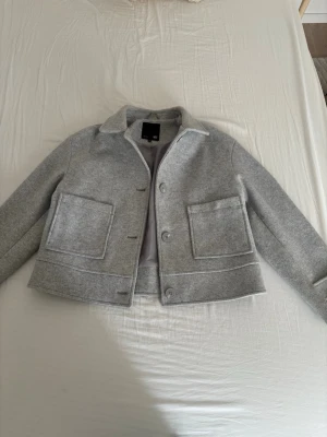 Grå kappa från Lager 157 - Snygg grå jacka från Lager 157 med stora fickor framtill och knappar. Jackan har en rak, croppad passform och klassisk krage. Perfekt till vardags för en stilren look.