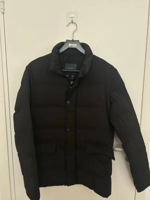 Svart pufferjacka från Pierre Cardin - Snygg svart pufferjacka från Pierre Cardin med vattenavvisande material och Futureflex-teknologi. Jackan har hög krage, knapp- och dragkedjestängning samt två stora fickor framtill. Perfekt för kalla vinterdagar och riktigt skön att bära.