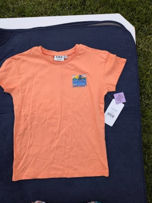 NY tshirt strl 122/128 - Snygg orange t-shirt från D.M.D med färgglatt strandmotiv och texten 'California Mavericks Beach' på ryggen. T-shirten har korta ärmar och är gjord i mjuk bomull, perfekt för en avslappnad stil.