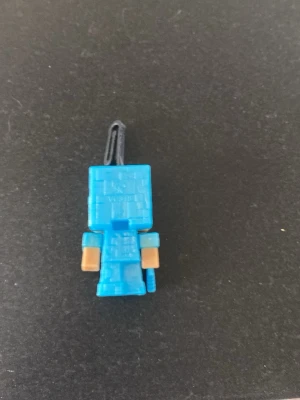 Blå Minecraft-figur nyckelring - Säljer en blå Minecraft-inspirerad figur som nyckelring. Tillverkad i plast med fyrkantig, pixlig design och beige detaljer på armarna. Perfekt att hänga på väskan eller nycklarna för en personlig touch. Från kinder joy.