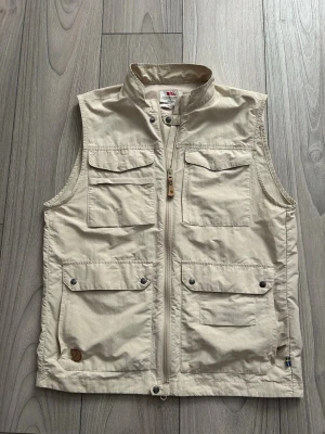 Beige väst från Fjällräven - Snygg beige väst från Fjällräven med fyra stora fickor framtill och dragkedja. Tillverkad i slitstarkt material och har diskret logga på ena fickan. Vid funderingar är det bara att höra av sig!
