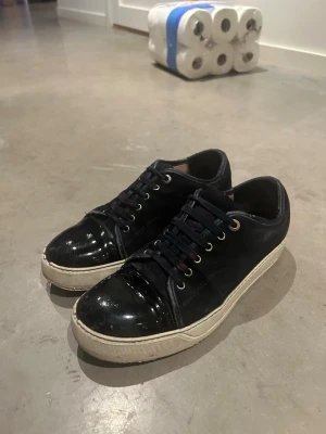 Lanvin - Stilrena svarta sneakers med glansig lackad tå och vit sula. Skorna har snörning med metallöljetter och en låg profil. Materialmix av textil och lack ger en modern vibe.