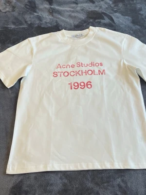 Vit Acne Studios t-shirt 1996 - Vit t-shirt från Acne Studios med röd text 'Acne Studios STOCKHOLM 1996' på bröstet. Klassisk passform med rund hals och korta ärmar. Perfekt för en clean och stilren look.