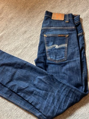 Mörkblå Nudie Jeans - Säljer ett par mörkblå jeans från Nudie Jeans med klassisk femficksmodell och kontrastsömmar. Vit nudie logga på bakfickorna, i nyskick knappt använda. Pågrund av att storleken är för kort för mig. Med mörkblå färg och lite nice ”slitningar”/mönster.  