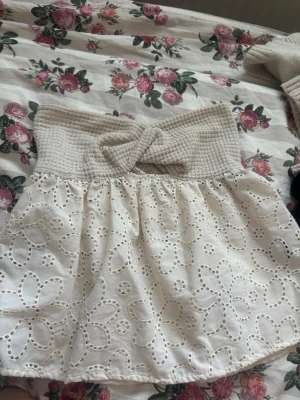 Vit broderad tubtopp med knut - Söt vit tubtopp med bred ribbad överdel och dekorativ knut framtill. Nederdelen är i broderad bomull med blommönster som ger en romantisk känsla. Perfekt för varma dagar och sommarens alla tillfällen.