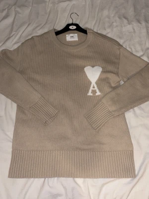 Beige stickad tröja från Ami Paris - Snygg beige stickad tröja från Ami Paris med rund halsringning och ribbade muddar. På bröstet finns det ikoniska vita hjärtat med bokstaven A. Tröjan har en rak passform och är tillverkad i mjukt material, perfekt för lager på lager. Pris kan diskuteras!