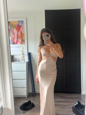 Lång champagnefärgad klänning - Säljer en elegant långklänning i champagnefärg med smala axelband och v-ringning. Klänningen har cut out-detaljer i midjan och figurnära passform. Materialet ser ut att vara satin eller silkesliknande för en lyxig känsla.