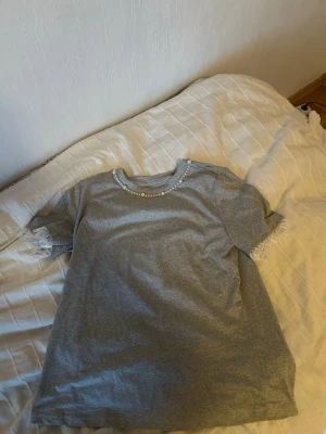 Grå t-shirt med pärldetaljer - Snygg grå t-shirt med korta ärmar och pärlor runt halsringningen. Ärmsluten har en liten spetskant som ger en extra touch. Perfekt för dig som vill ha något enkelt men ändå med lite detaljer.