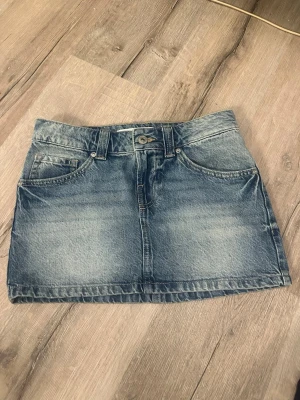Blå jeanskjol med fickor - Snygg blå jeanskjol i klassisk modell med fem fickor, bälteshällor och knappstängning fram. Kjolen har en kort passform och är tillverkad i slitstarkt denimtyg. Perfekt att matcha med en t-shirt eller hoodie för en avslappnad look.