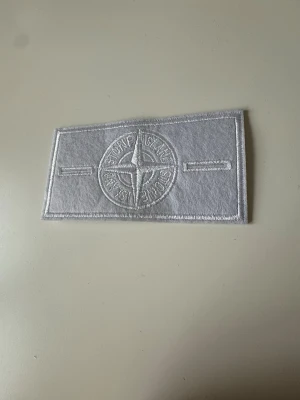 Stone island badge - white - Säljer ett vitt tygmärke från Stone Island med broderad kompasslogga. Märket är rektangulärt och har detaljer i vitt, perfekt att sy eller fästa på jackor, väskor eller andra plagg för en unik look.